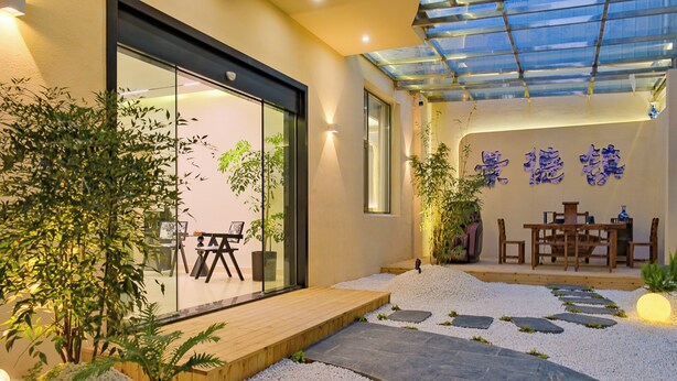 Ein moderner Außenbereich mit Terrassenbereich, Glastür, Holzdeck und einem Kiesweg, der zu einem Essbereich mit Tisch und Stühlen führt.