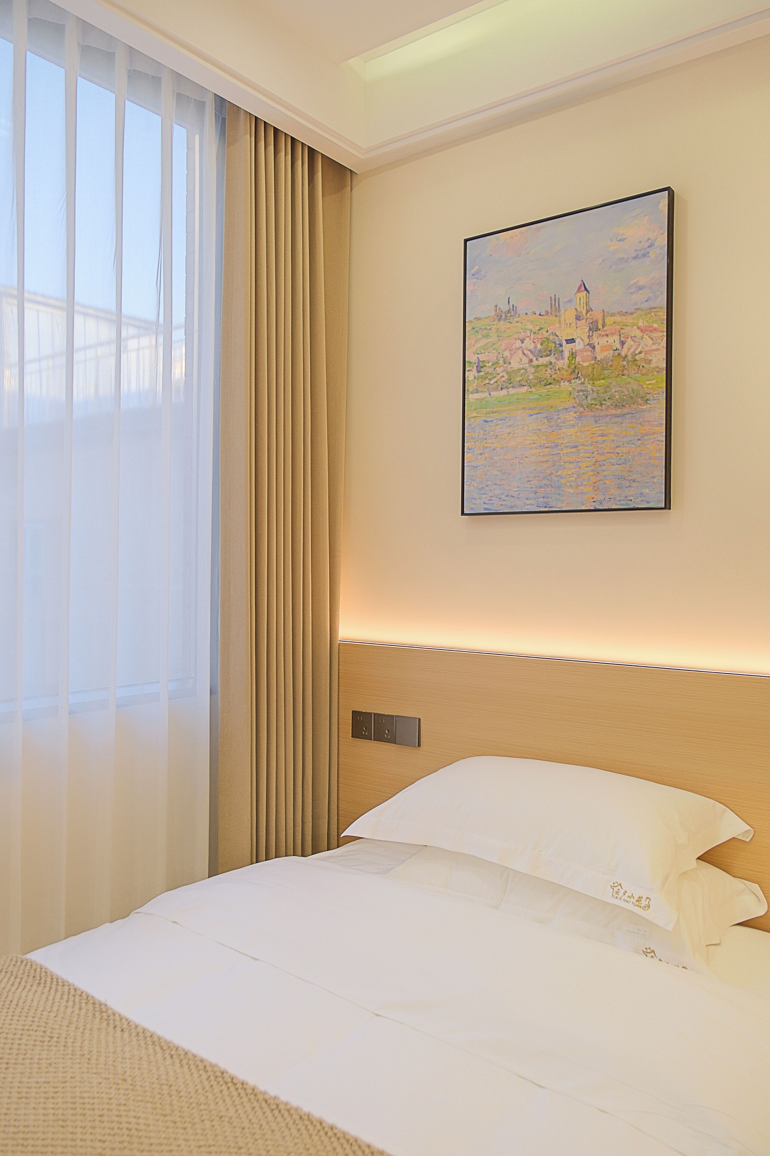 A hotel room with a bed, a painting of a landscape, and a window with curtains.