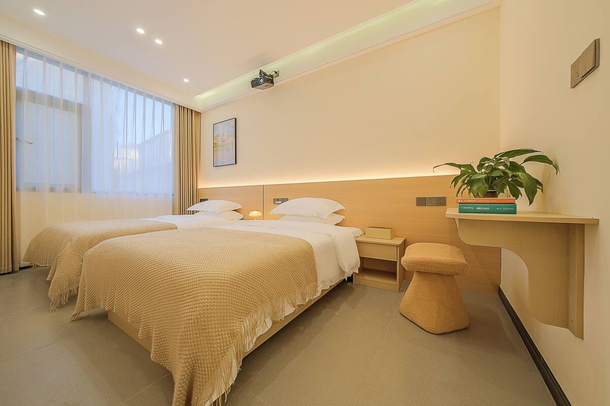 A hotel room with two beds, a bedside table with a lamp, a small stool, a shelf with a plant, and a projector on the ceiling.