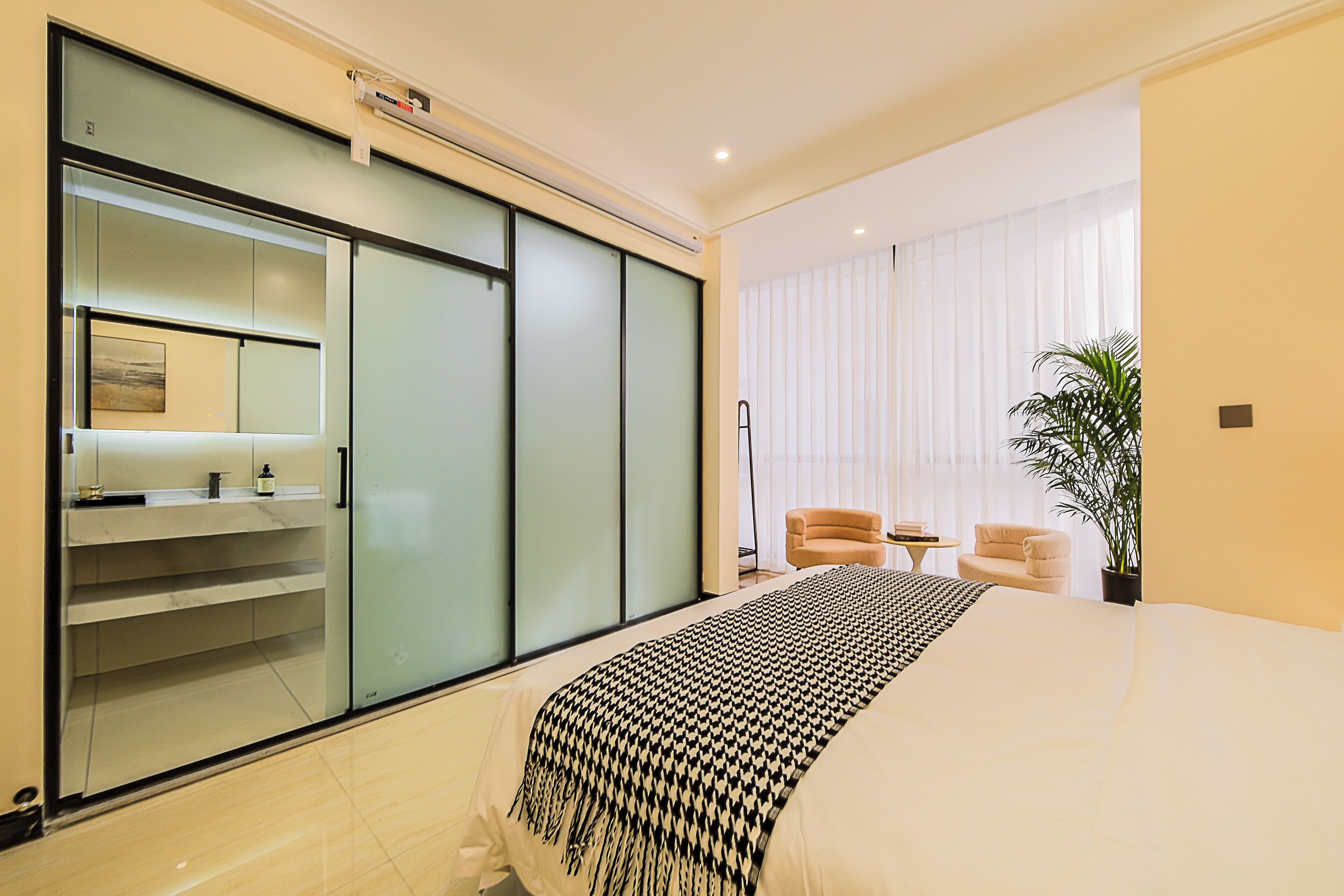 Ett modernt hotellrum med en stor säng, en skjutdörrsgarderob i glas, en liten sittgrupp med två stolar och ett stort fönster med tunna, genomskinliga gardiner.