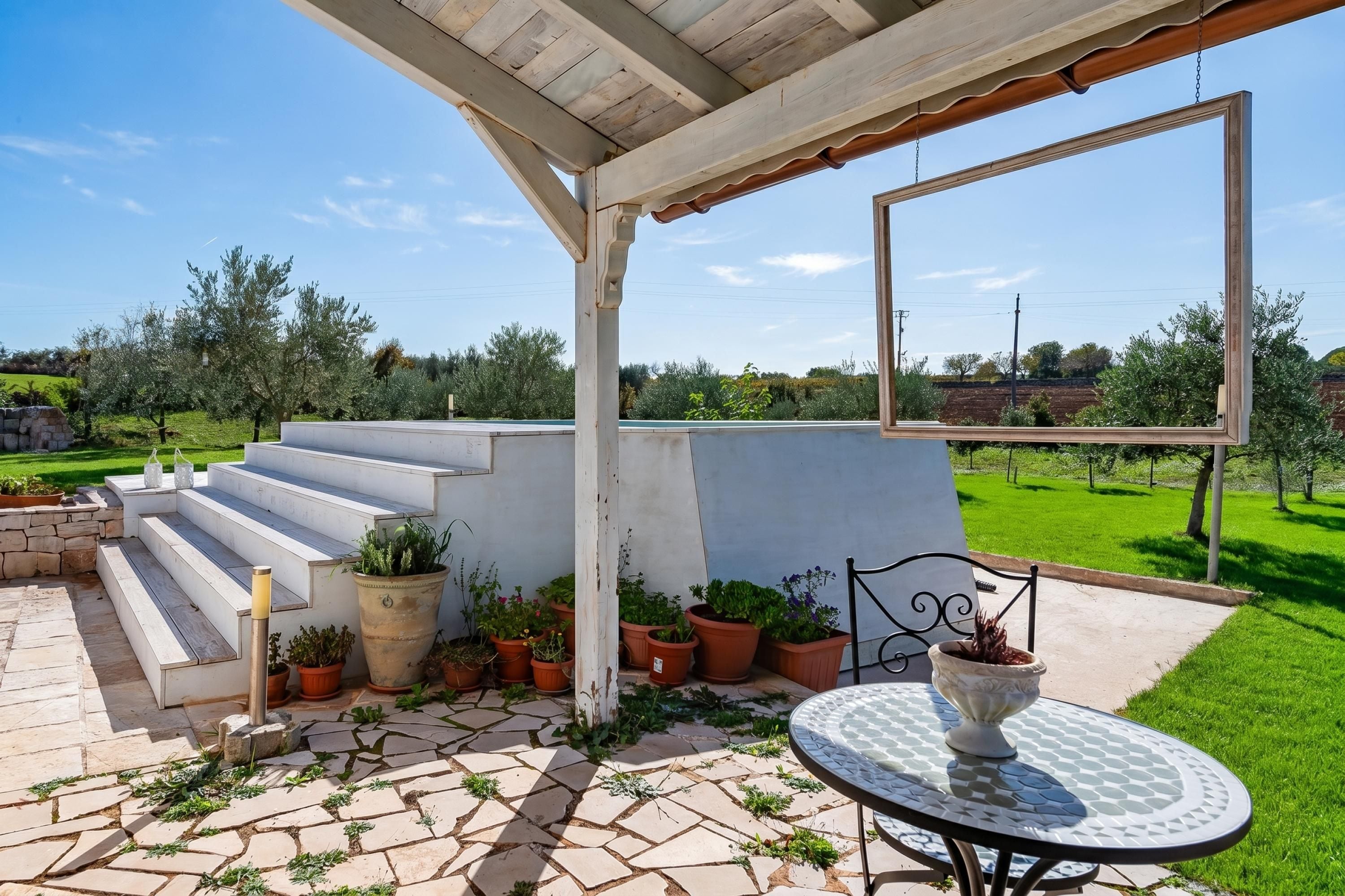 A covered patio with steps leading to a white wall, potted plants, and a table with a bowl.