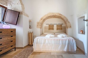 A bedroom with a stone arch, a wooden dresser, a bed with white linens, and a mounted television. - Trullo Conosolo With Pool (Martina Franca)