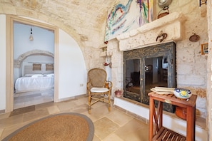A room with a bed, a wicker chair, a fireplace, and a small table with a bowl. - Trullo Conosolo With Pool (Martina Franca)