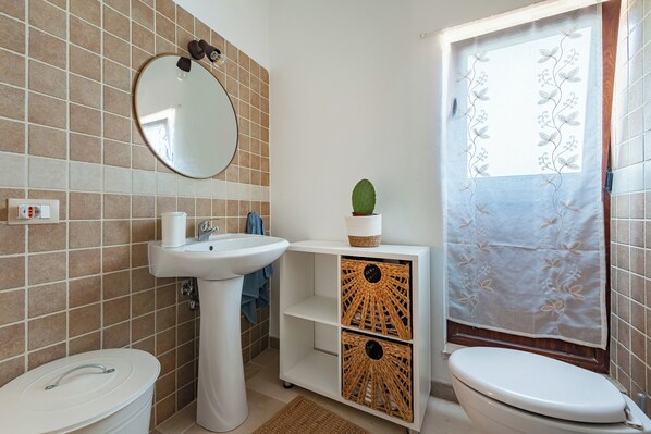 A bathroom with a white sink, toilet, and a mirror. There is a plant on the shelf and a shower curtain with a floral pattern. - Trullo Conosolo With Pool (Martina Franca)