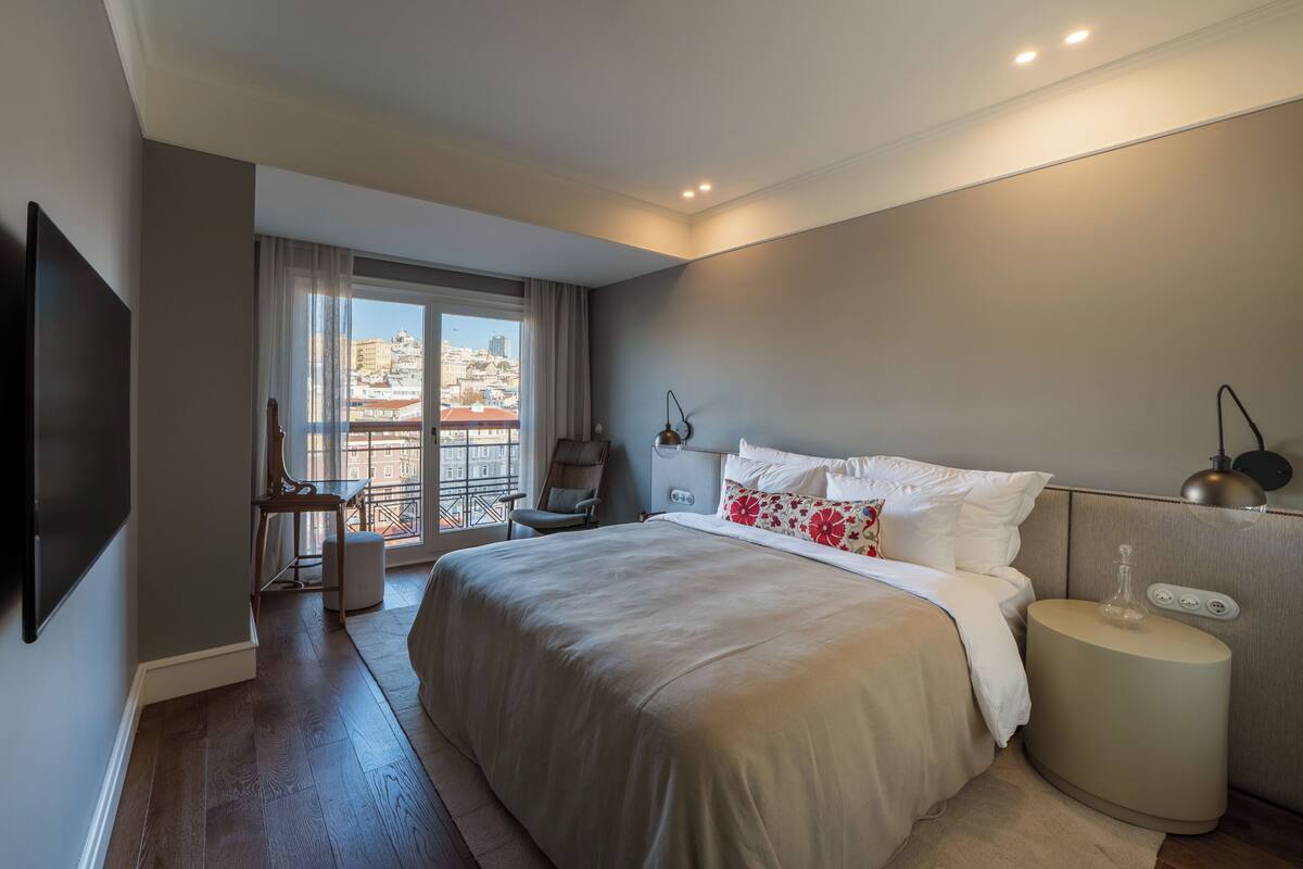 A modern hotel room with a large bed, a desk, a chair, a TV, and a balcony with a city view.