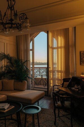 A room with a sofa, a dining table, a chair, a potted plant, and a view of the water outside.