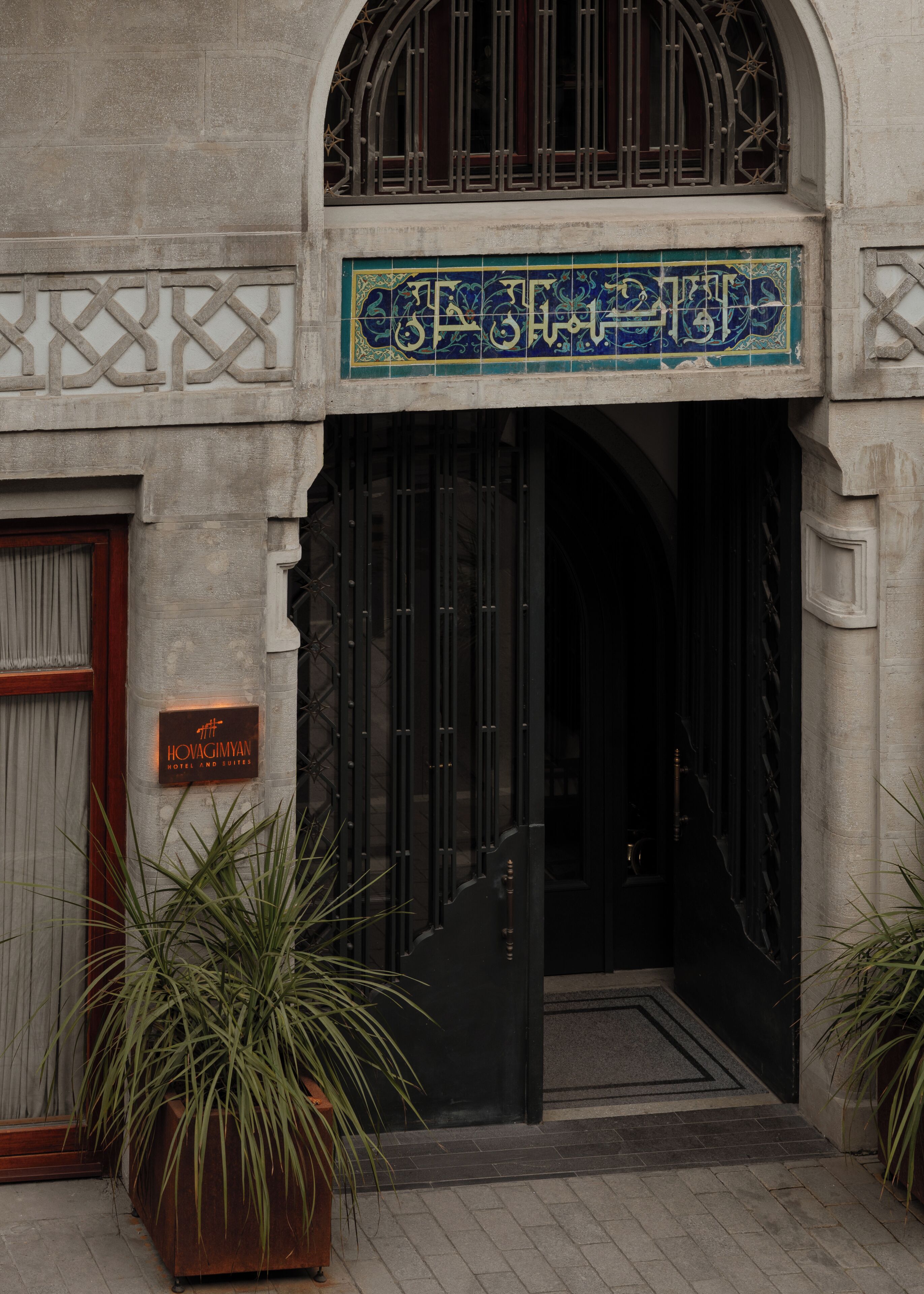 A hotel entrance with a sign reading 'HOVAGIMYAN HOTEL AND SUITES,' a black gate, and potted plants.