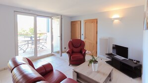 A modern living room with a red leather sofa, a red armchair, a wooden TV stand, and a sliding glass door leading to an outdoor patio with a table and chairs. - Entre Plages & Espagne : Ferme de Charme, Pistes cyclables & Baignade Bio (Orx)