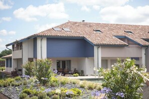 Ein modernes Haus mit Ziegeldach, einer überdachten Terrasse und einem von einem Garten mit verschiedenen Pflanzen und Blumen umgebenen Swimmingpool.