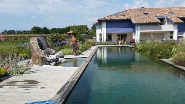 A swimming pool with a wooden deck, a house with a blue garage door, and a person walking by. - Entre Plages & Espagne : Ferme de Charme, Pistes cyclables & Baignade Bio (Orx)