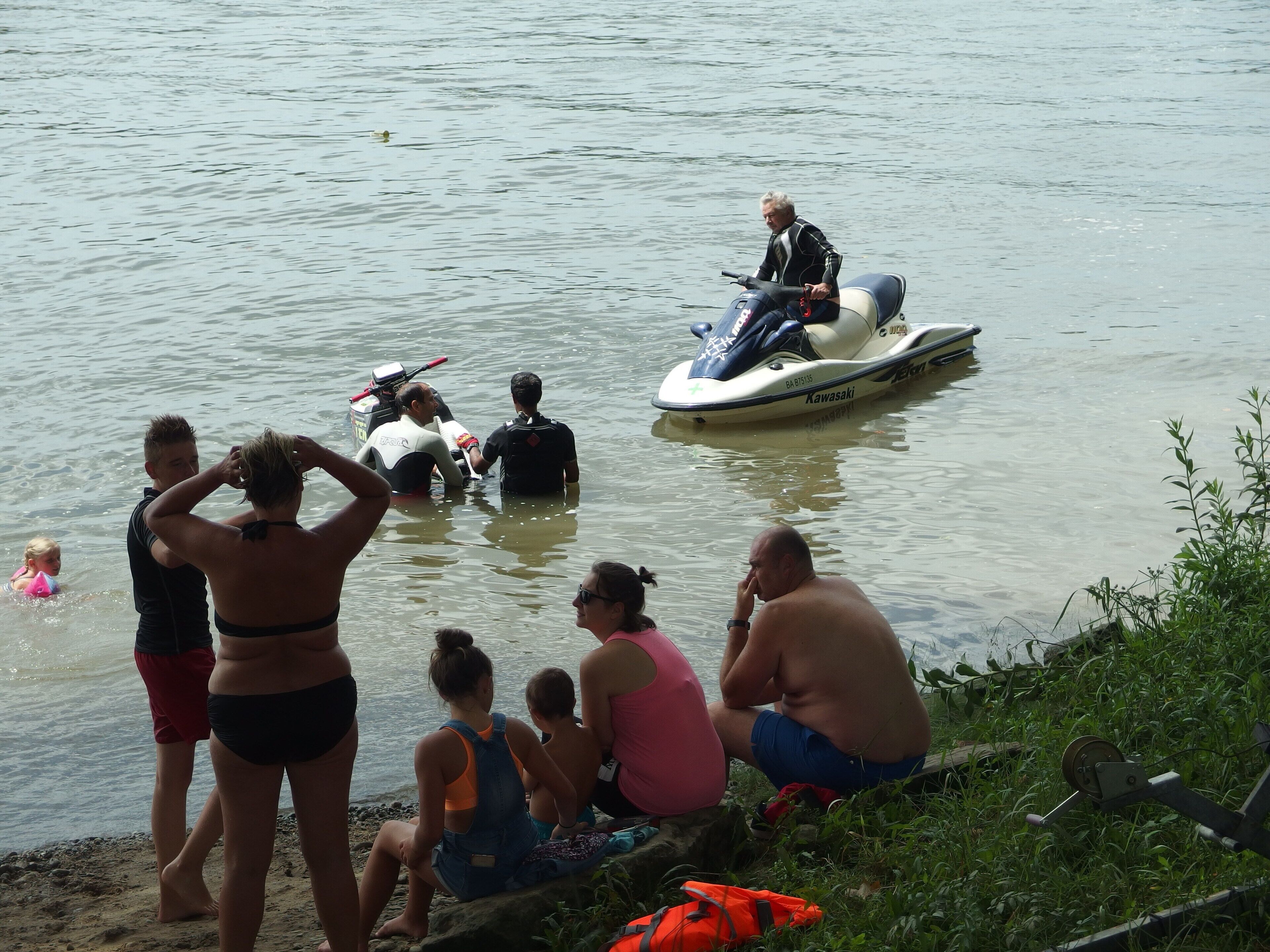Een groep mensen bij een meer, sommigen in het water en anderen aan de oever.