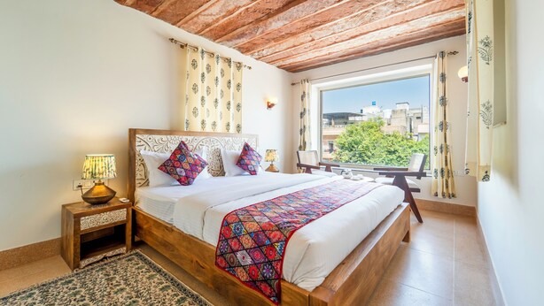 A bedroom with a wooden bed, patterned curtains, a view of buildings, and a tiled floor. - The Munshi Mansion (Jodhpur)