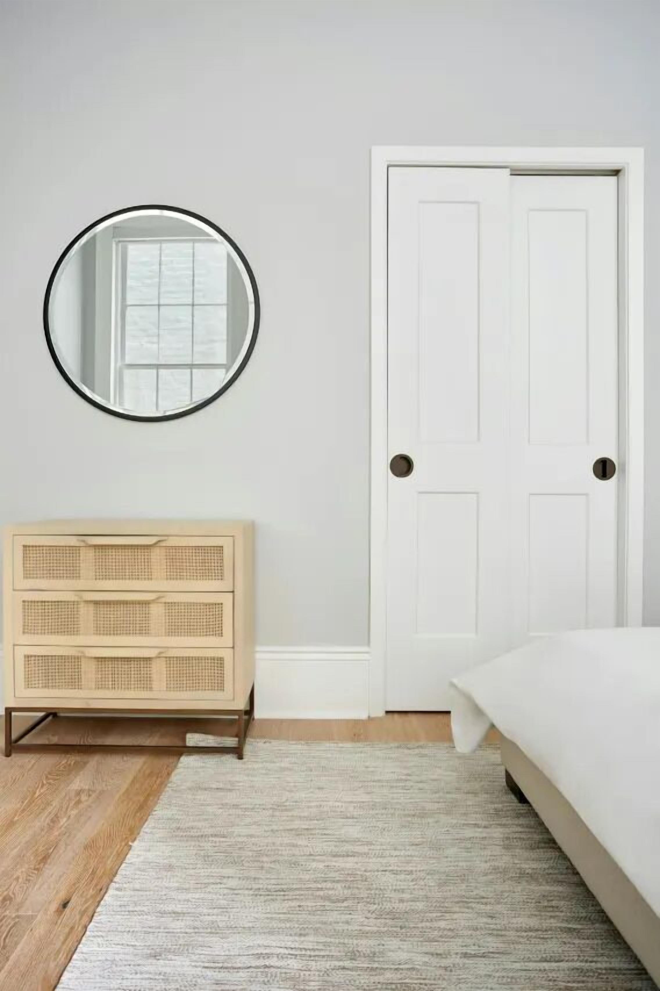 Un dormitorio con una cama, un aparador de madera, un espejo redondo y una puerta blanca.