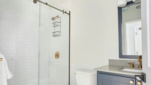 Ein modernes Badezimmer mit Duschkabine aus Glas, weißen Fliesen, einer weißen Toilette und einem dunkelblauen Schrank mit goldenem Wasserhahn.