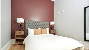 A bedroom with a tufted headboard, a large bed, two bedside tables with lamps, and a wooden nightstand. - Steps to Bourbon Street | Hearing-Accessible 2BDR (New Orleans)