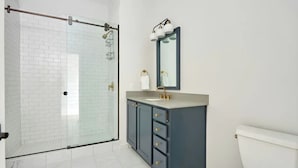 A modern bathroom with a glass-enclosed shower, a dark vanity with gold handles, and a white toilet. - Steps to Bourbon Street | Hearing-Accessible 2BDR (New Orleans)