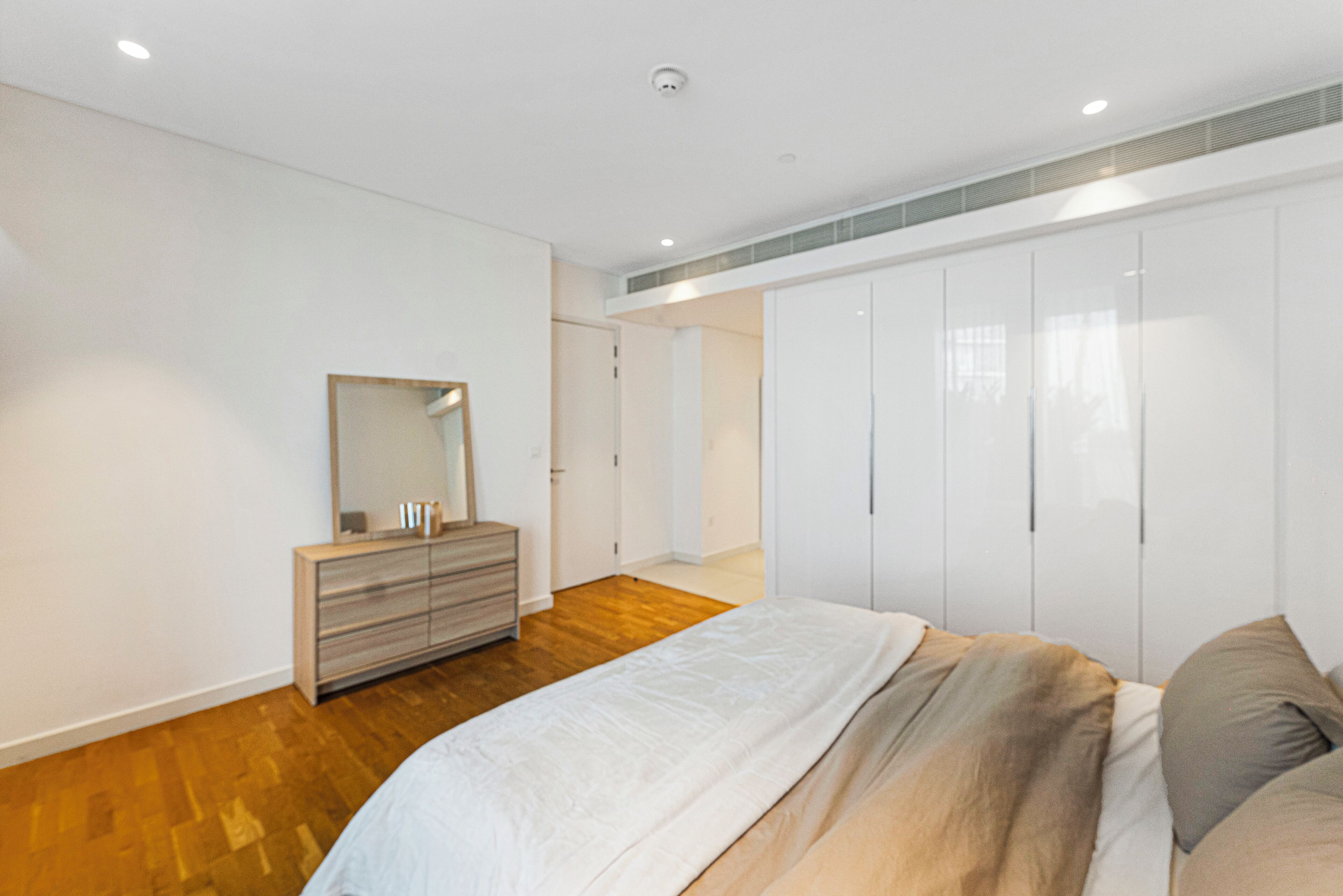 Une chambre avec un plancher en bois, un lit recouvert d’une couette blanche et beige, une commode avec un miroir et une armoire aux portes blanches.