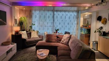 A modern living room with a brown sofa, a small wooden coffee table, and a lamp. There is a window with sheer curtains and a view of the outside.