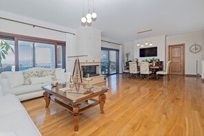 A spacious living room with a white sofa, a wooden coffee table, and a fireplace. There is a dining area with a table and chairs, and a large window offering a view of the outdoors.