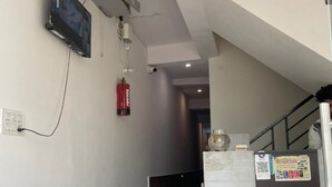 A hotel room with a bed, a small wooden bench, a TV mounted on the wall, a fire extinguisher, and a refrigerator with a poster. - Devi Heritage Vrindavan (Vrindavan)