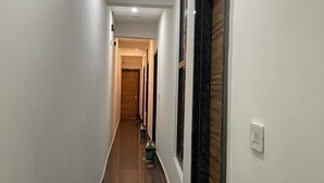 A long, narrow hallway with white walls and a glossy black floor. There are several doors on the right side, one of which is open. A trash bin is visible on the floor. - Devi Heritage Vrindavan (Vrindavan)
