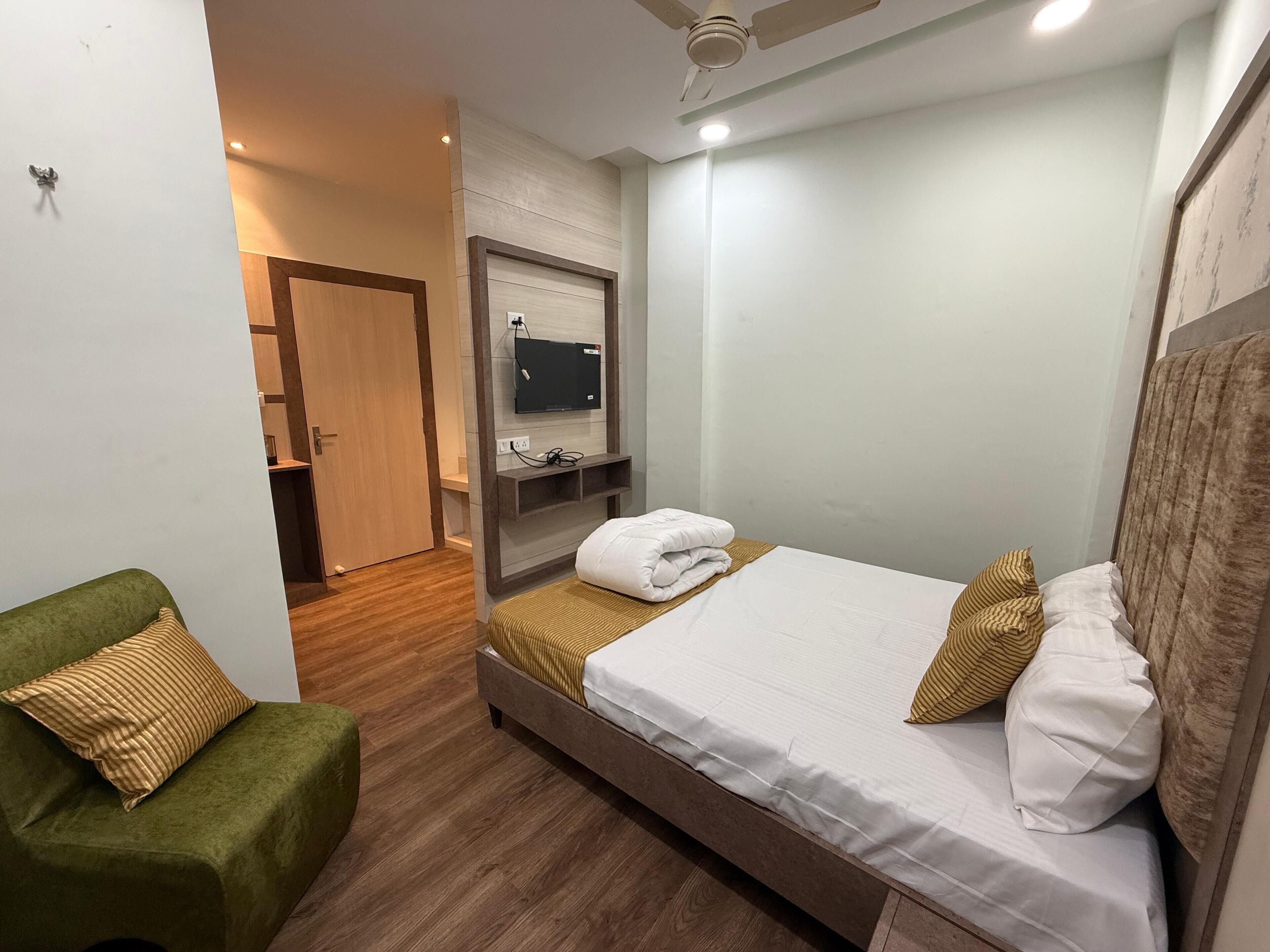 Une chambre d’hôtel avec un lit, un fauteuil vert, un téléviseur fixé au mur et un plancher en bois.