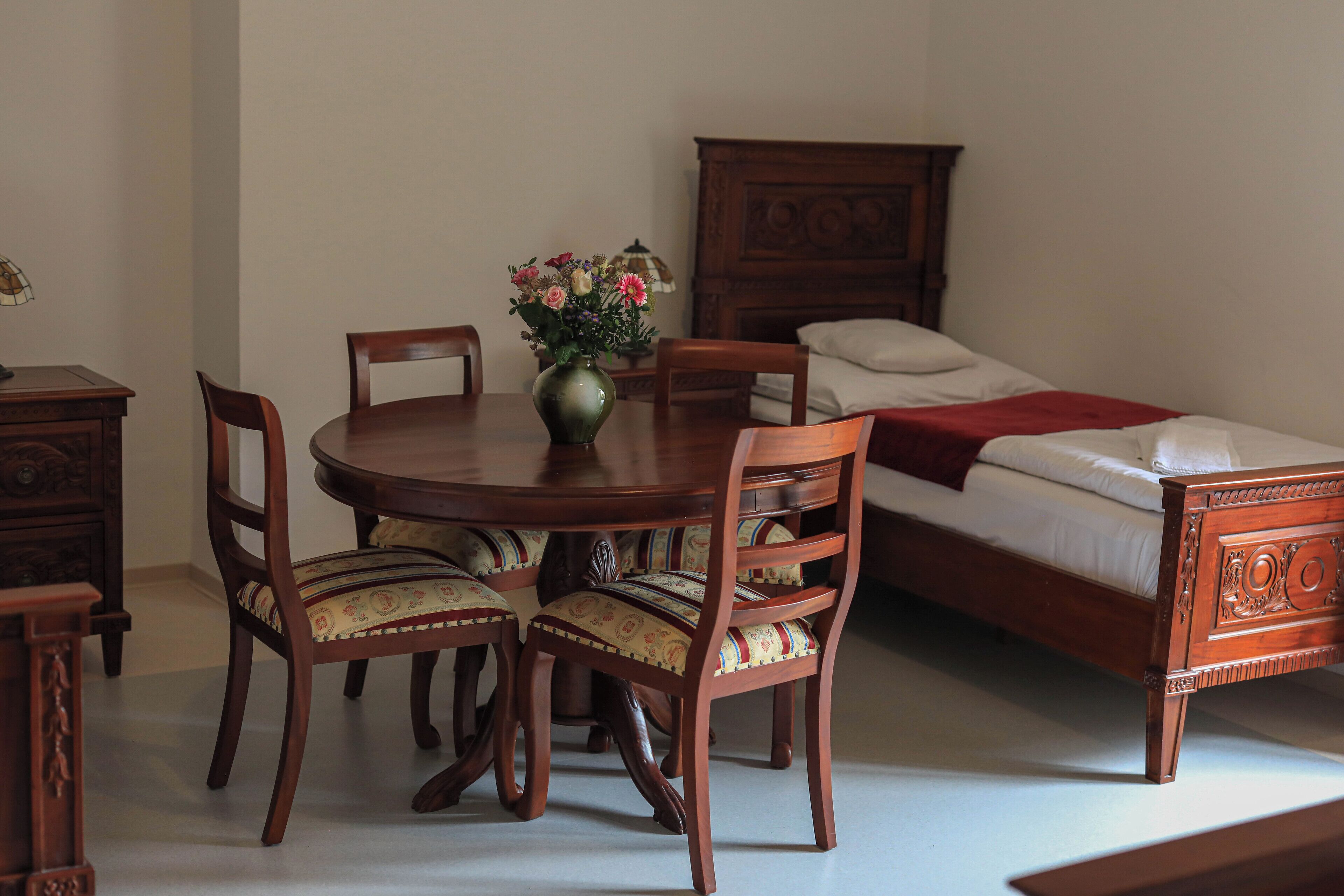 A room with a wooden dining table, four chairs, a vase with flowers, a bed with a wooden headboard, and a nightstand.