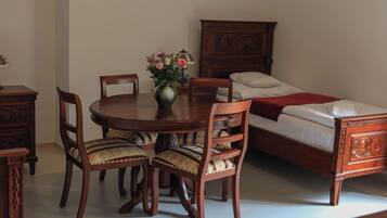 A room with a wooden dining table, four chairs, a vase with flowers, a bed with a wooden headboard, and a nightstand.
