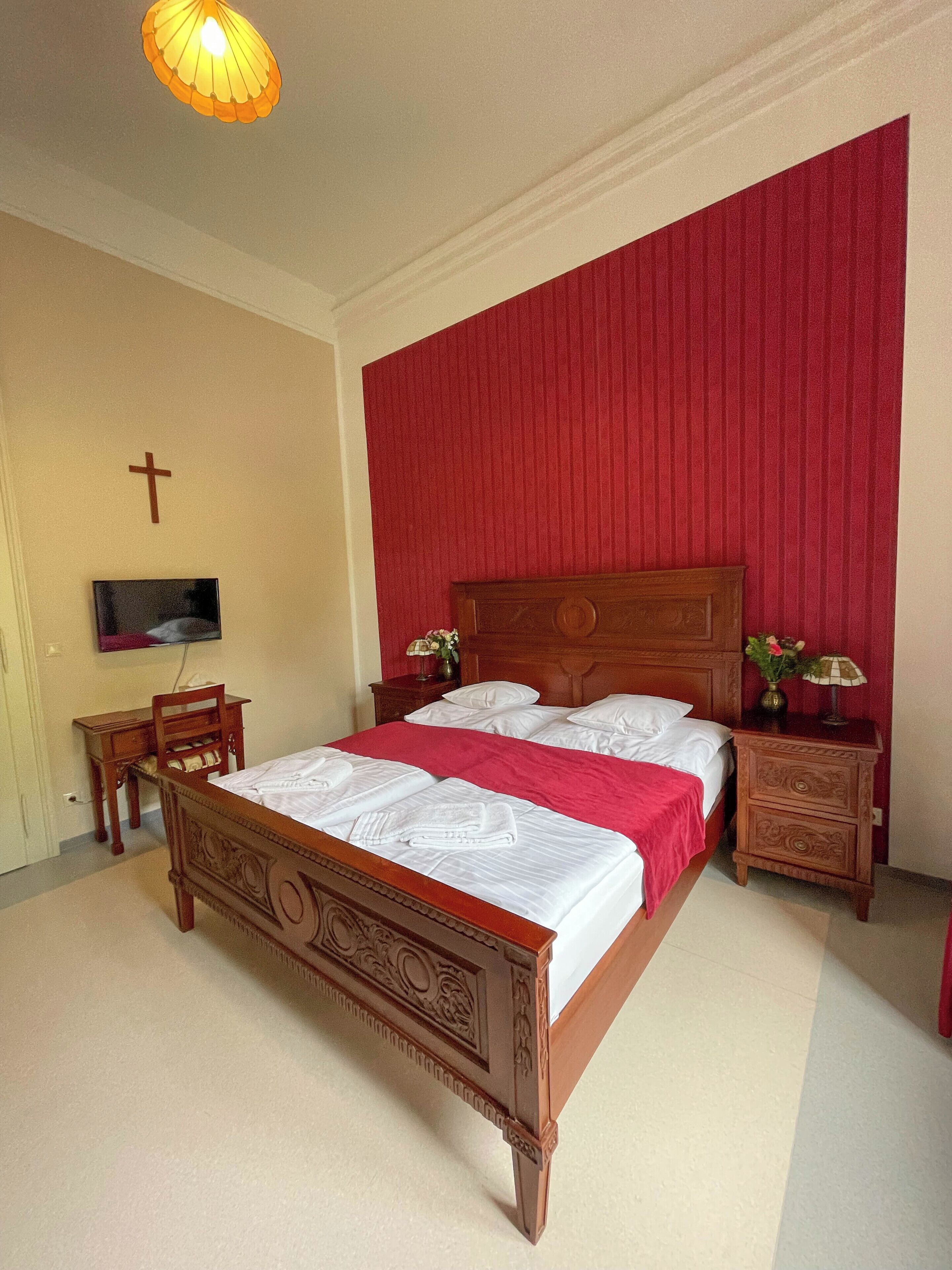A neatly arranged bedroom with a wooden bed, a television on the wall, and a cross decoration.