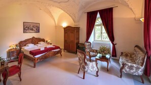 Ein Zimmer mit einem großen Bett, zwei Sesseln, einem Holzschrank und einem Fenster mit roten Vorhängen.