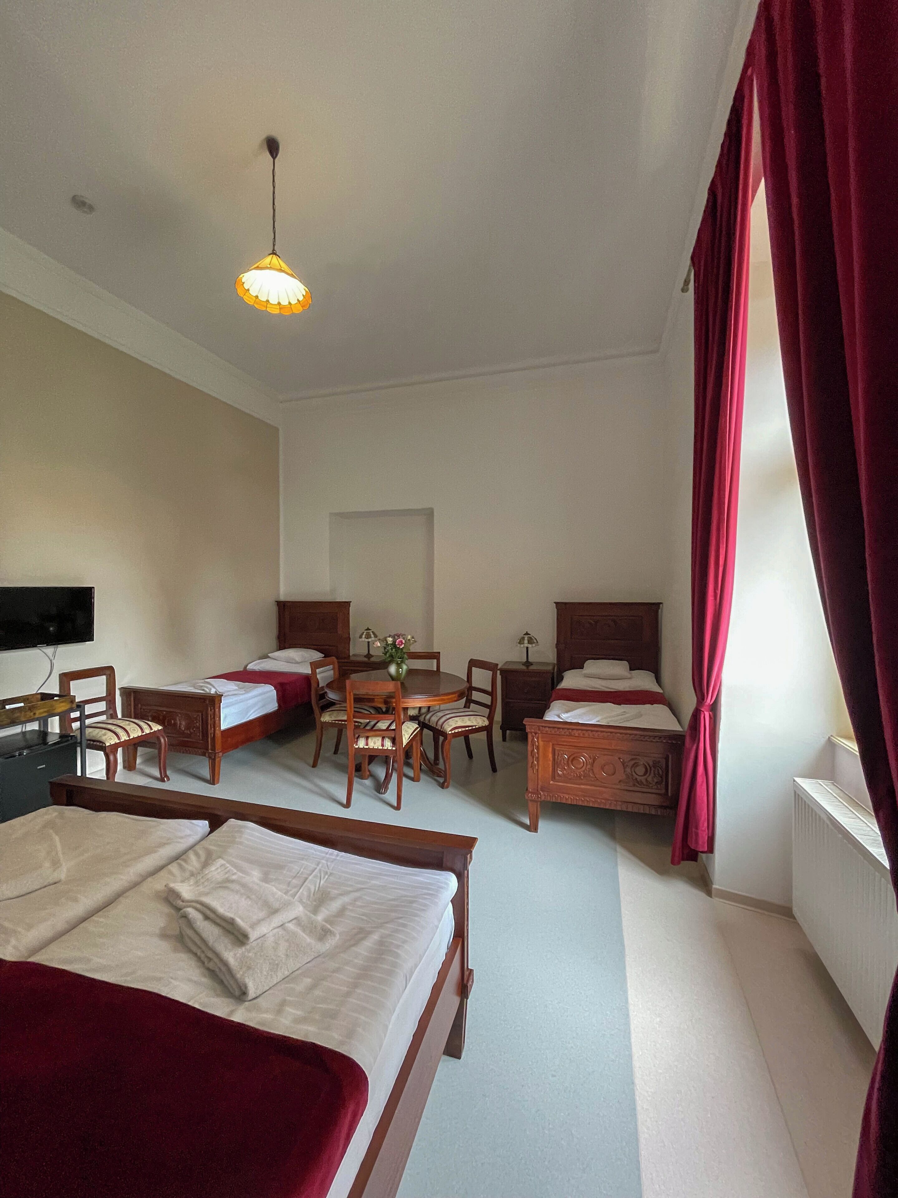 Une chambre d’hôtel avec deux lits simples, un bureau, une chaise, une table ornée d’un vase de fleurs et une fenêtre avec des rideaux rouges.