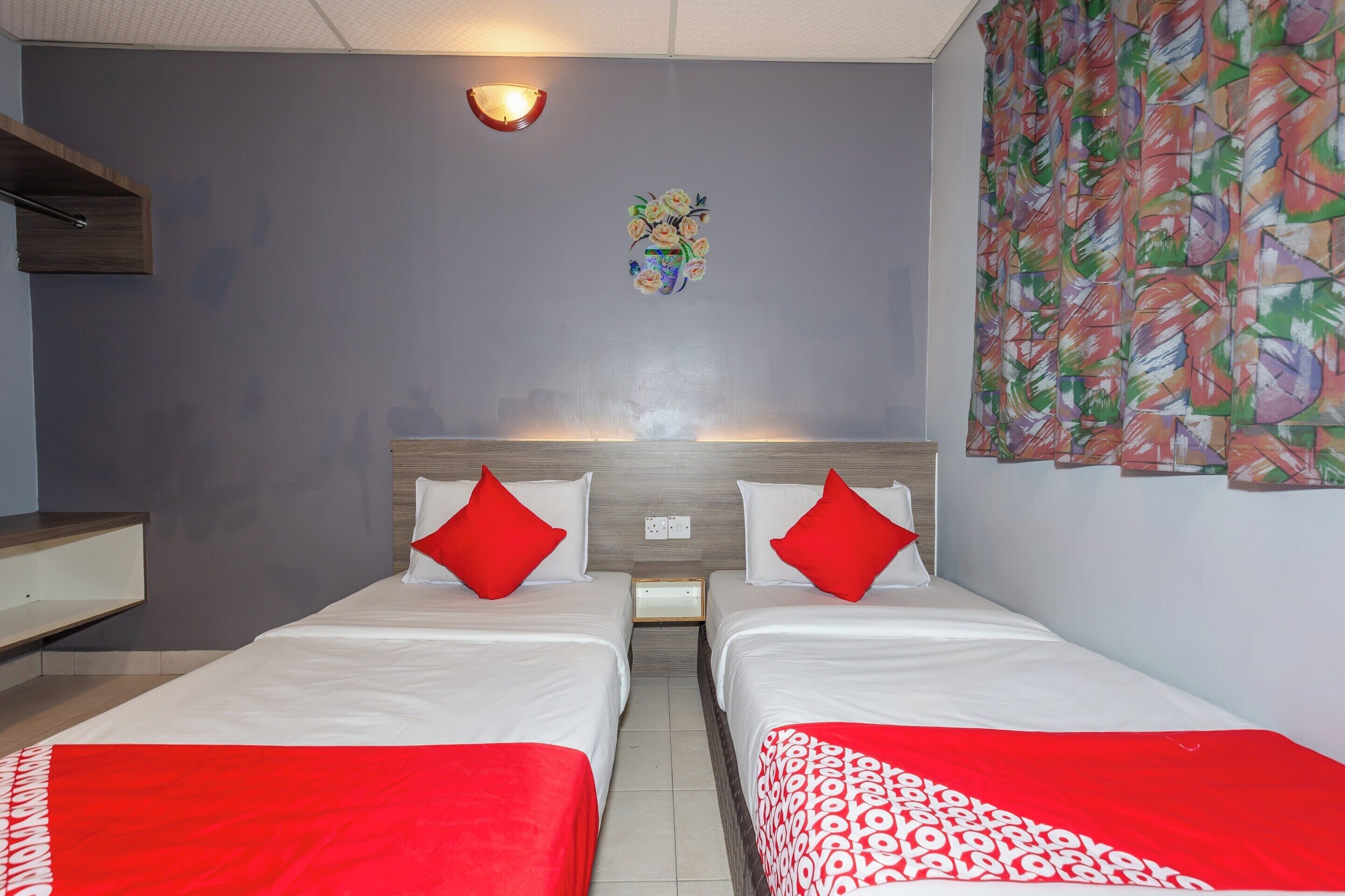 Habitación de hotel con dos camas, ropa de cama roja y blanca, una mesita de noche, una lámpara de pared y un cuadro abstracto de colores.