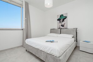 Ein modernes Schlafzimmer mit einem grauen Bett, weißer Bettwäsche, einem Nachttisch mit einem Buch und einem Bild einer Frau mit grünem Hut an der Wand.