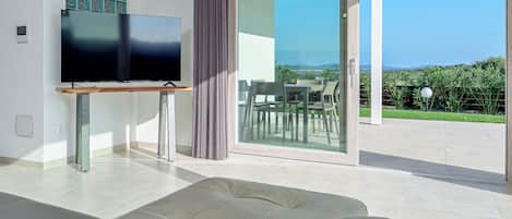 A modern living room with a flat-screen TV, a sofa, and a sliding glass door leading to an outdoor patio with a table and chairs.