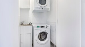 Ein kleiner Waschbereich mit einer Frontlader-Waschmaschine und einem Toplader-Trockner, beide weiß, mit einem Edelstahlspülbecken und einer Bedienblende an der Waschmaschine.