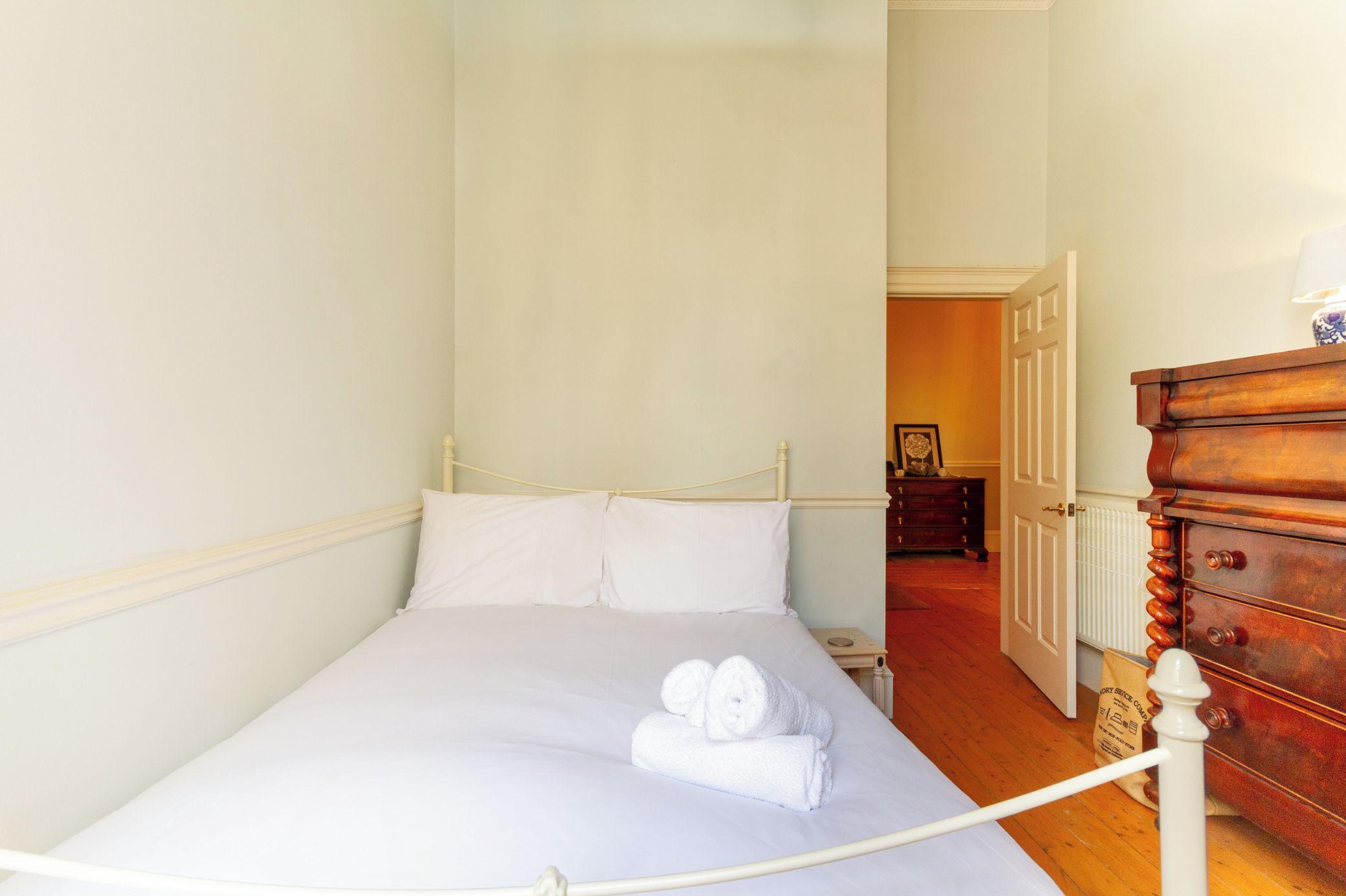 Una cama bien hecha con sábanas blancas y dos toallas enrolladas, un aparador de madera con cajones, y una puerta que da a otra habitación.