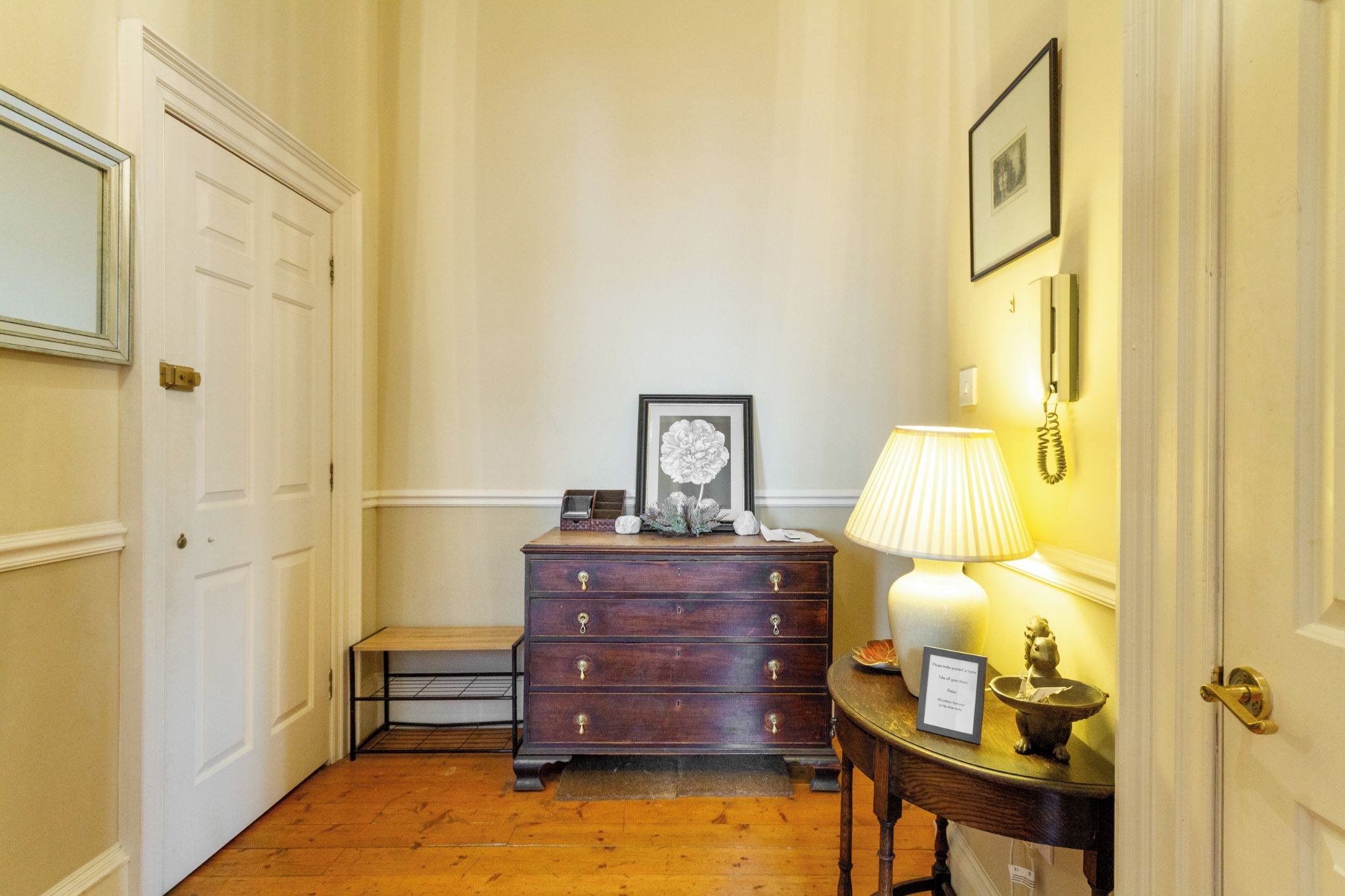 A hallway with a wooden floor, a dark wooden dresser with drawers, a small table with a lamp, and a framed picture on the wall.