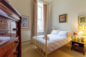 Ein Schlafzimmer mit einem Bett, einem Schminktisch, einem Fenster mit Vorhängen, einer Heizung und gerahmten Bildern an der Wand.