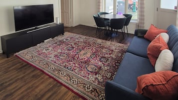 A living room with a flat-screen TV, dark sofa, patterned rug, dining table, and chairs.