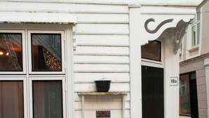 Un bâtiment blanc avec une porte noire portant l’inscription « 10a ». On y trouve deux chaises en bois et une petite table à l’extérieur.