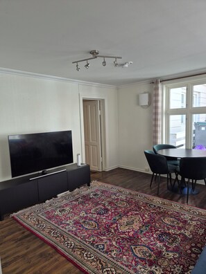 Ein Wohnzimmer mit Flachbildfernseher, einem dunklen Sofa, einem gemusterten Teppich, einem Esstisch mit Stühlen und einem Fenster mit Vorhängen.
