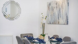 Ein Essbereich mit einem Glastisch, grau gepolsterten Stühlen, einem dekorativen Spiegel und einem abstrakten Kunstwerk an der Wand.