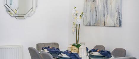 A dining area with a glass table, grey upholstered chairs, a decorative mirror, and a piece of abstract art on the wall.