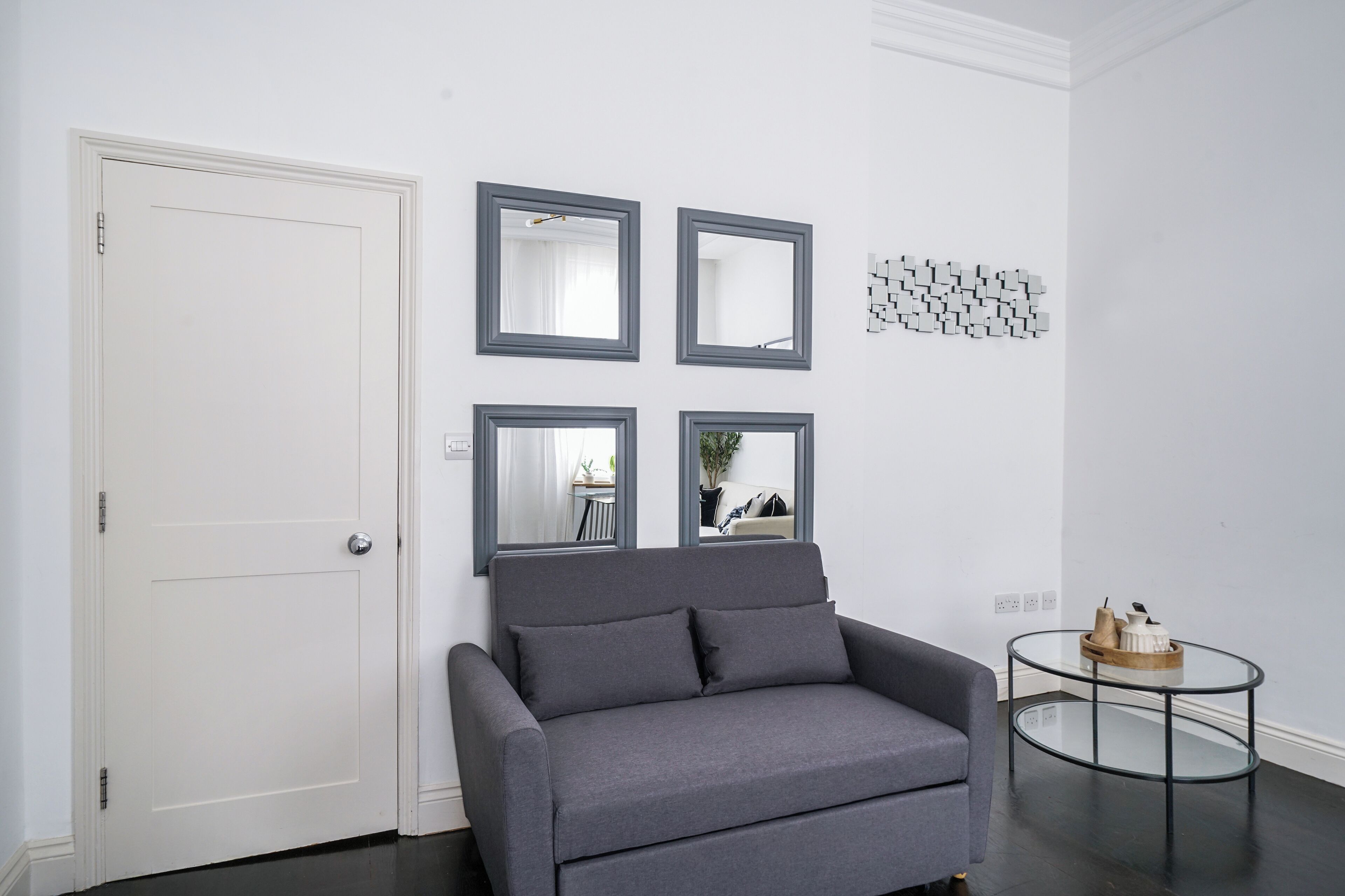 Een kamer met een grijze bank, een witte deur en een glazen salontafel met een houten dienblad.