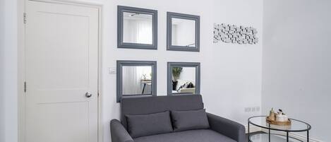 Een kamer met een grijze bank, een witte deur en een glazen salontafel met een houten dienblad.