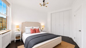 Ein Schlafzimmer mit Bett, Nachttischen, Heizkörper, großem Kleiderschrank und Fenster mit Blick auf Gebäude.