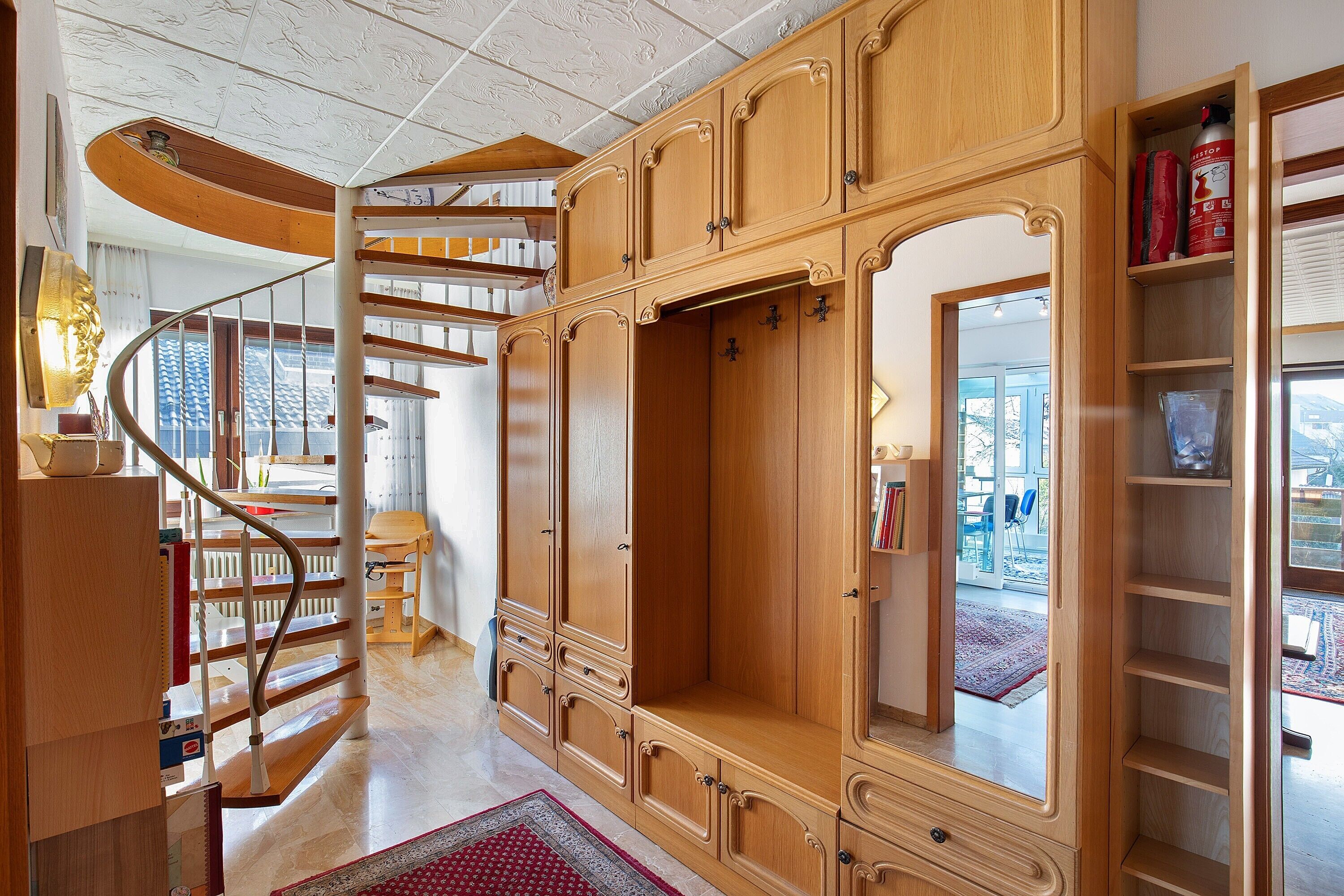 Eine Wendeltreppe aus Holz führt zu einer oberen Etage, ein großer Holzschrank mit mehreren Türen sowie ein Bücherregal mit Büchern und Dekorationsobjekten.