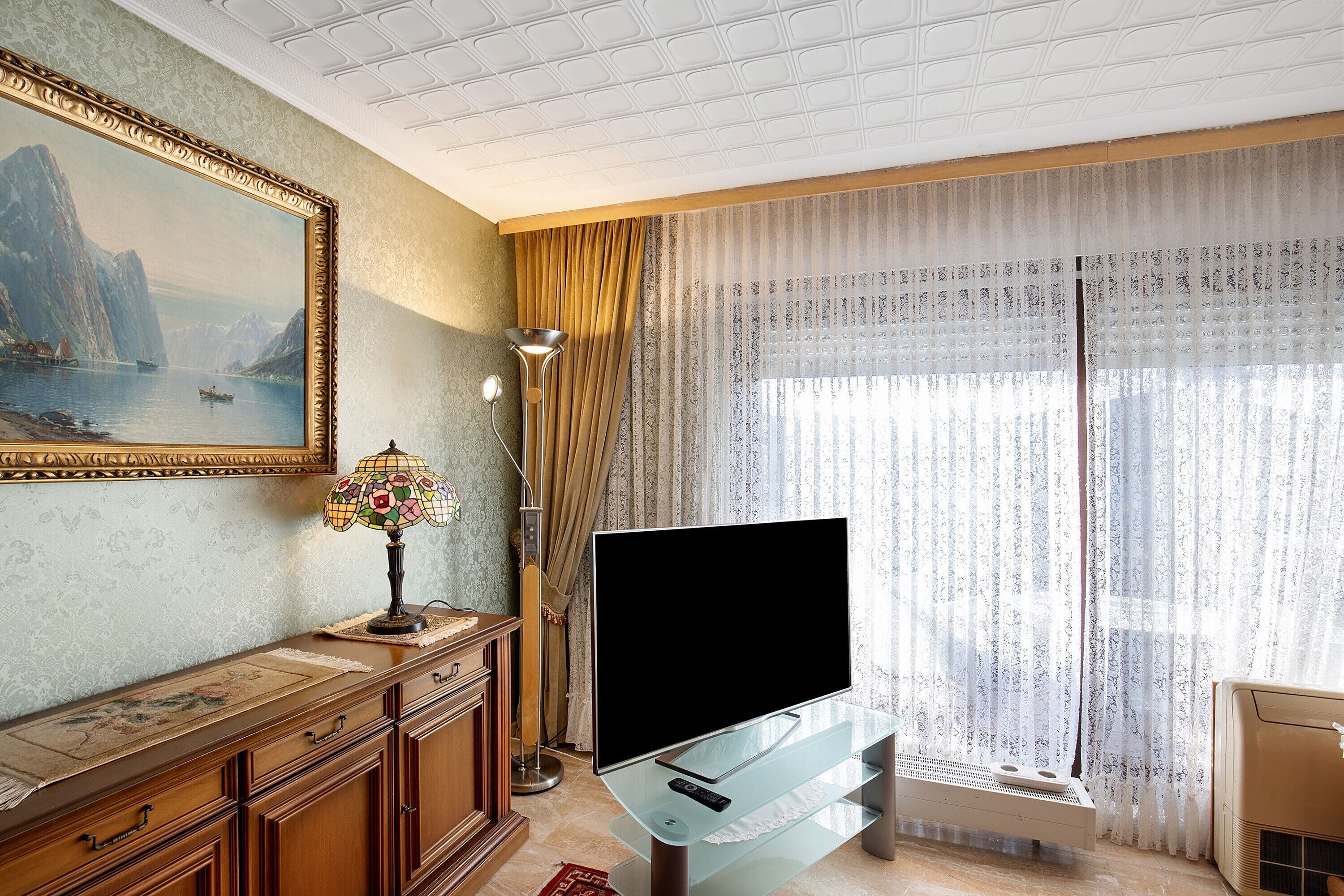 A room with a flat-screen TV on a glass stand, a wooden cabinet with drawers, a framed painting, a floor lamp, and sheer curtains with a view of the outdoors.