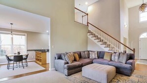 Ein geräumiges Wohnzimmer mit Ecksofa, Hocker und einem Essbereich mit Tisch und Stühlen. Eine Treppe mit Geländer führt zu einer oberen Ebene.