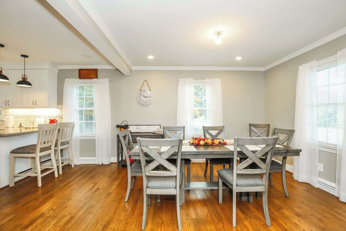A dining area with a wooden table and chairs, a clock on the wall, and a window with curtains.
